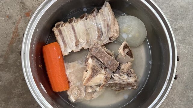 Metal pot with boiled pork bones, carrot and onion in water on concrete floor. Homemade broth, soup cooking, bone meal, pet food preparation