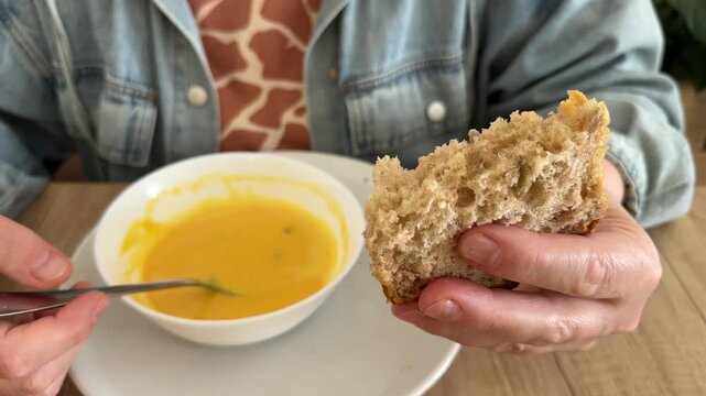 Person holding piece of whole grain bread and spoon over bowl of pumpkin soup at wooden table. Healthy lunch, homemade meal, vegetarian food
