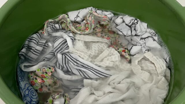 White and patterned clothes soaking in soapy water in green plastic basin. Hand wash, laundry day, household chores, cleaning at home