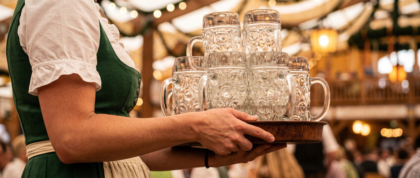 Woman server carrying a tray of traditional beer mugs inside a festive Bavarian beer hall. Oktoberfest celebration, hospitality service, German culture, food festival atmosphere, and event catering