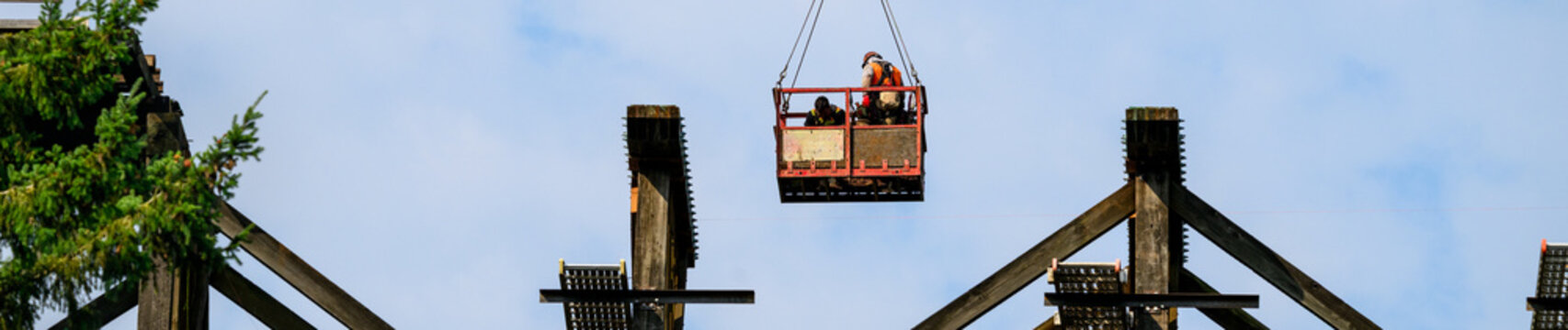 Construction workers in arial work basket over old tall wooden railroad trestle, train tracks removed, structural framework update, sunny summer day, America
