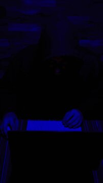 Hooded hacker typing in dark blue lit room performing cyber attack and data breach concept