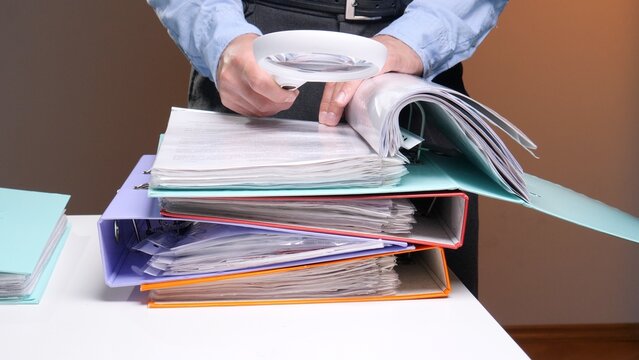 Man in a blue shirt carefully examining financial papers in a binder with a magnifying glass, performing an audit or detailed investigation of corporate reports and legal compliance documents
