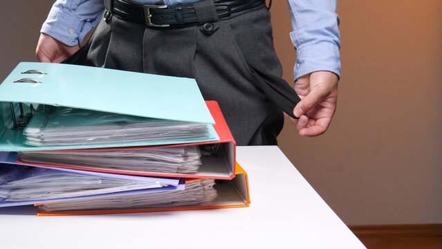 Person standing behind a stack of binders and documents, turning out pants pockets to show they are empty, symbolizing financial problems and a lack of money despite hard work