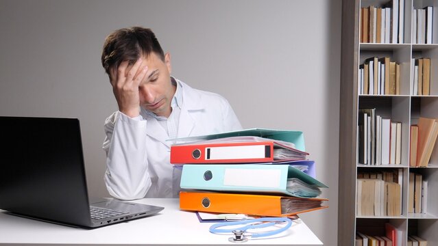 Exhausted healthcare professional sitting at a desk with a laptop and a large stack of colorful binders, holding head in hand, indicating stress and being overworked