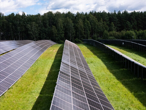 Aerial view of rows of solar panels installed in a green field bordered by a dense forest under a cloudy sky in Kaunas, Kaunas County, Lithuania.