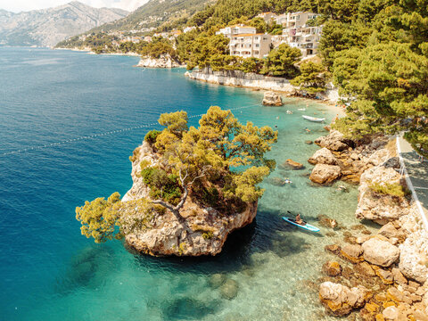 Aerial view of the iconic Brela Rock surrounded by turquoise Adriatic Sea waters, with a kayaker paddling nearby, lush pine trees, and coastal houses in Brela, Split-Dalmatia County, Croatia.
