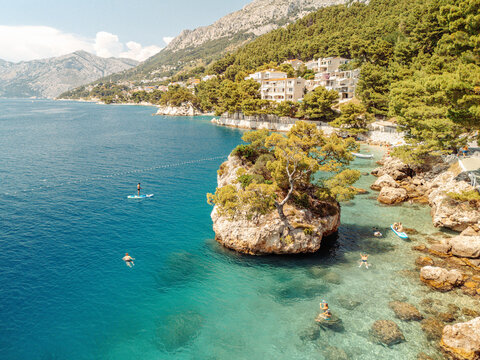 Aerial view of the iconic Brela Stone rock formation in turquoise Adriatic water, with people swimming and paddleboarding near the rocky coastline in Brela, Split-Dalmatia County, Croatia.
