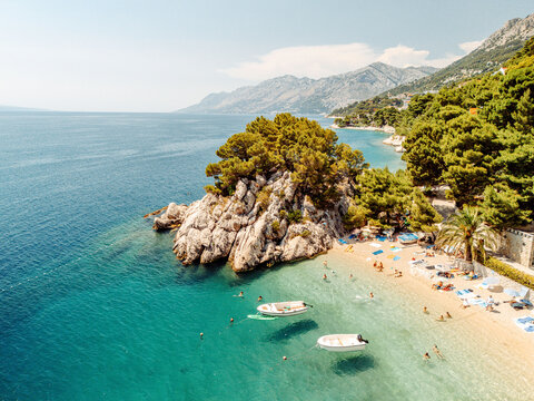 Aerial view of the iconic Brela beach with its distinctive rock formation, lush pine trees, turquoise water, and small boats anchored near the shore in Brela, Split-Dalmatia County, Croatia.
