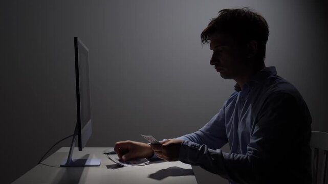 Man in silhouette sitting at a desk, counting cash money next to a computer monitor, representing illegal online activity, cybercrime, fraud, and dark web transactions