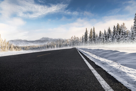 A straight empty asphalt road cuts through a snowy coniferous forest landscape on a clear cold winter day, with distant mountains under blue sky.