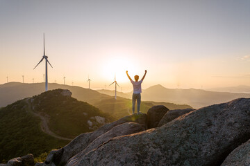 A person stands on a mountain peak, raising arms to celebrate sunrise with wind turbines in the scenic outdoor landscape. © tai