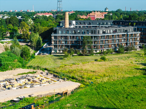 Aerial view of a modern residential apartment building with a brick chimney, situated next to a construction site with green lawns and trees in Riga, Latvia.