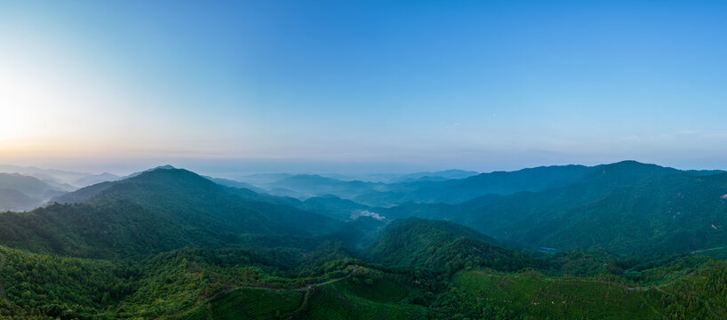 Panoramic view of layered green mountain ranges at dawn under a clear blue sky, with mist in valleys.