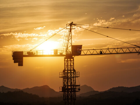 A tower crane silhouette stands against a golden sunset sky with distant mountains, capturing industrial construction at dusk.