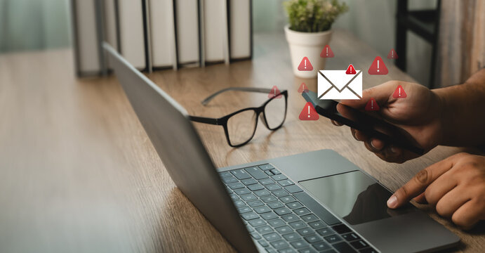 Hands using a smartphone beside a laptop with email envelope and warning icons, showing mail and digital communication alerts, message notifications online inbox monitoring.