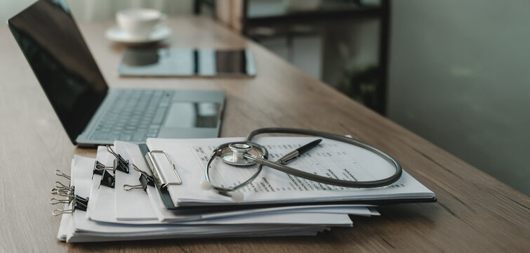 Stethoscope rests on printed documents beside laptop and pen on wooden desk, with clipboard and paper stack arranged for medical documentation, healthcare records, and clinical management.
