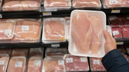 Fototapeta premium Person holding packaged raw chicken breast in supermarket aisle among meat products on display. concept of grocery shopping, food selection, fresh poultry, consumer choice, meal preparation