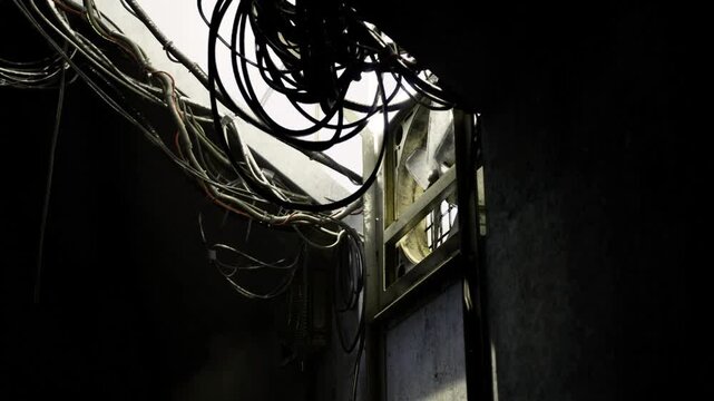 Overhead cable tangle near window, daylight filtering through grime, exposed conduit and fraying insulation suggest urgent repair, hazardous wiring and confined