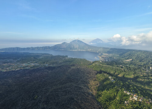 Aerial view of Mount Abang and Lake Batur surrounded by volcanic lava fields and lush green vegetation under a clear blue sky in Kintamani, Bali, Indonesia.