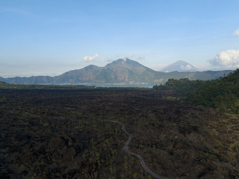 Aerial view of Mount Abang rising above the vast volcanic lava fields with a winding road cutting through the rugged terrain in Kintamani, Bali, Indonesia.