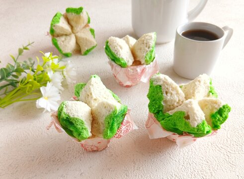 Traditional Indonesian steamed sponge cake, known as bolu kukus, with a distinctive flower-like shape and vibrant green and white colors, served in paper cups with a cup of black coffee. Soft, fluffy 