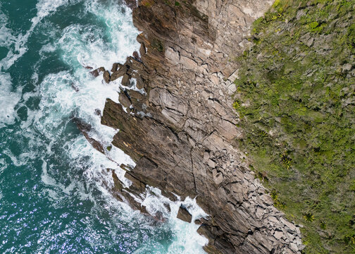 Aerial view of Praia da Foca featuring jagged rocky cliffs meeting the turquoise ocean with white crashing waves and lush coastal vegetation in Armacao dos Buzios, Rio de Janeiro, Brazil.
