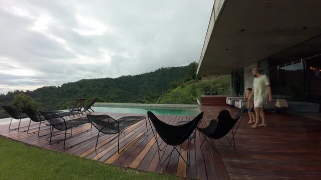 FPV drone shot of luxury concrete villa in Costa Rica with infinity pool wooden deck and rainforest hills as father and son walk together past lounge chairs under misty tropical sky.