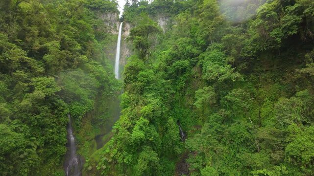 FPV aerial flight through a misty rainforest canyon in Costa Rica revealing a tall waterfall plunging over a cliff into dense green jungle with tree ferns and vines on steep wet slopes.