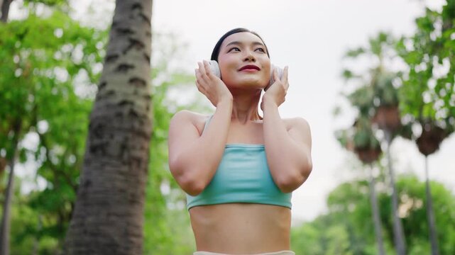 Asian woman wearing wireless headphones and activewear, standing in a green public park enjoying fresh air with a relaxed and refreshed expression. Outdoor fitness and wellness lifestyle concept