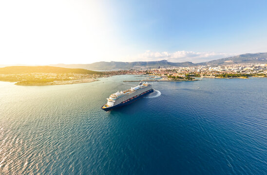 Aerial view of a large cruise ship sailing into the Port of Split, with the historic city skyline and coastal mountains illuminated by golden hour sunlight, Split, Split-Dalmatia County, Croatia.