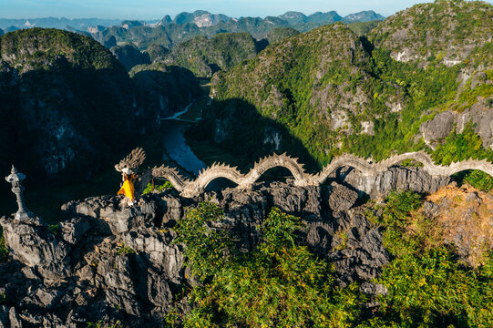 Aerial view of the dragon statue atop the rocky cliffs of Mua cave with a person in a yellow dress standing nearby, overlooking the landscape of Nam Hoa Lu, Ninh Binh, Vietnam.