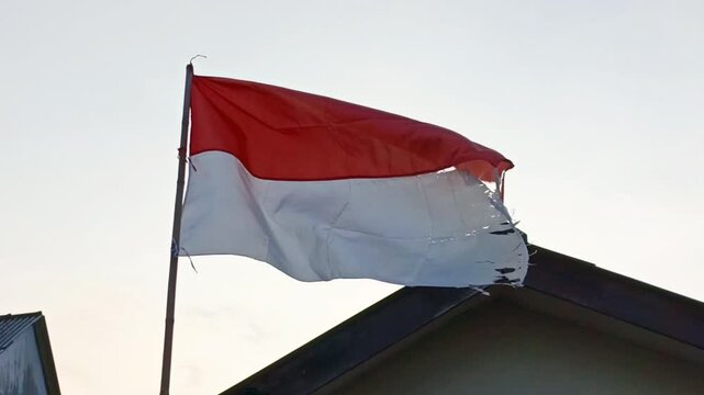 Footage of a torn Indonesian flag waving on a pole above a rooftop under clear sky.