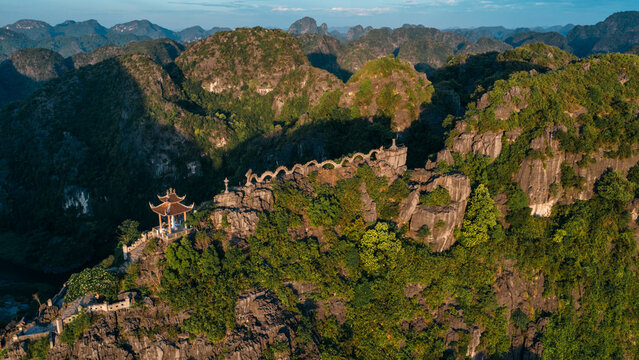 Aerial view of Mua Cave with its iconic dragon statue on the limestone mountain ridge, surrounded by lush green karst landscape under golden hour light in Nam Hoa Lu, Ninh Binh, Vietnam.