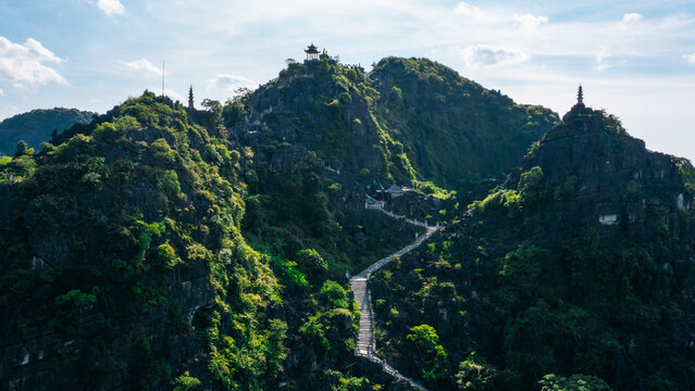 Aerial view of Mua Cave with its iconic stone staircase winding up the lush green karst mountain, topped by a pagoda under a bright blue sky in Nam Hoa Lu, Ninh Binh, Vietnam.