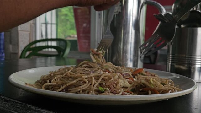 Freshly prepared stir-fried noodles onto a plate at a local Indian restaurant or roadside food establishment. The scene shows noodles being lifted with utensil, highlighting everyday food preparation