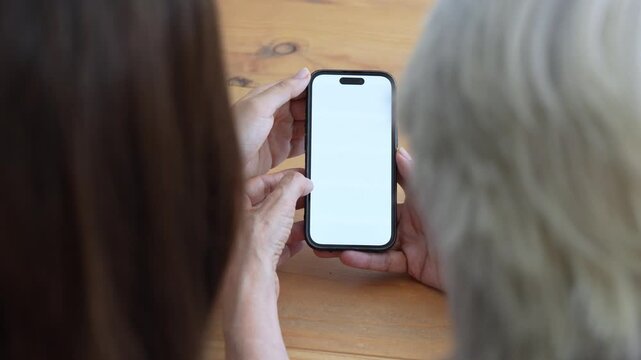 High-angle view of a daughter teaching her elderly mother how to use a new mobile phone, showing her how to scroll and tap on the white blank screen while sitting together at a wooden table