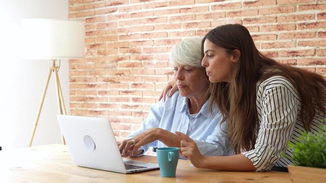 Supportive young woman helping her senior grandmother use a laptop, explaining how to navigate the internet and pointing at the screen while the elder woman types and learns