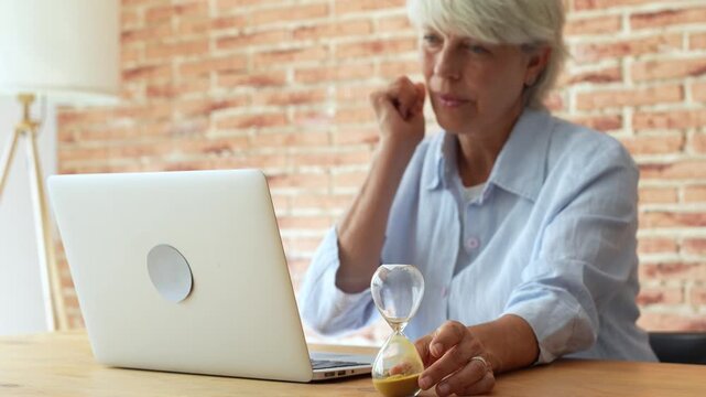 Elderly woman with grey hair working on her laptop computer at a desk, turning over an hourglass to measure time, symbolizing productivity, deadlines, and the passing of time in business