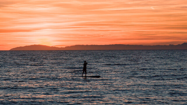Aerial view of a person paddleboarding on the calm ocean during a vibrant orange sunset with distant coastline in Laguna Beach, California, United States.