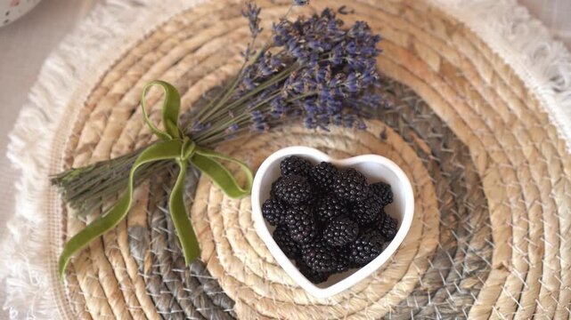 Top view of black raspberry berries blackberries in white porcelain heart shaped plate on natural round herbal napkin with dried bouquet of lavender tied velvet green ribbon