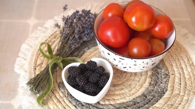 Bowl of ripe tomatoes and heart-shaped dish of blackberries arranged on a woven placemat with dried lavender, creating a rustic and inviting kitchen scene 