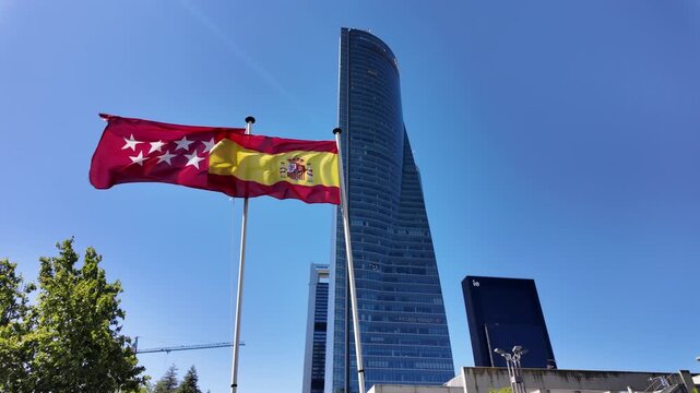 Close view of the Spanish and Community of Madrid flags moving in the wind with a tall glass tower behind them.