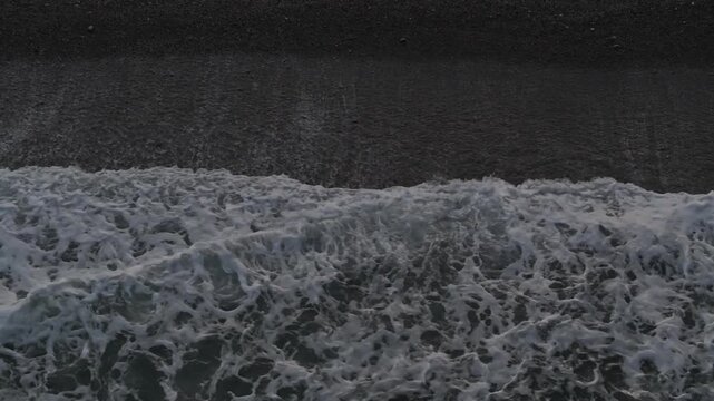 Overhead top down aerial shot of sea waves crashing and creating white foam water patterns on dark sandy beach shore during calm daylight, ideal for background texture or nature concept.