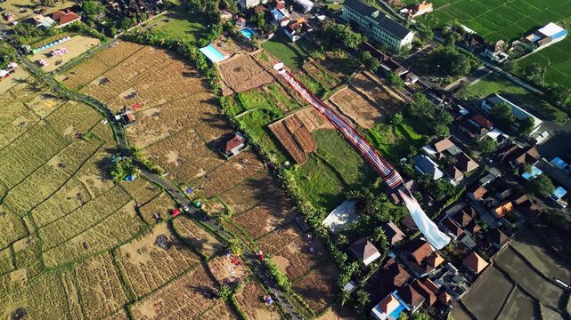 Traditional Balinese kite flies above green rice fields in Bali Indonesia revealing geometric farmland patterns village homes and tropical vegetation overhead drone footage