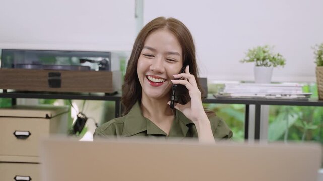 Happy beautiful Asian woman sitting smiling and talking on her cell phone with customer about work with laptop on the desk in office. Pretty young female freelance using mobile phone in the workplace.