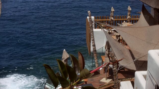 Terrace in Costa Adeje, Tenerife, sits above waves breaking against the rocky coast. High-angle view shows wooden decking, shade sails, closed parasols, and oceanfront seating