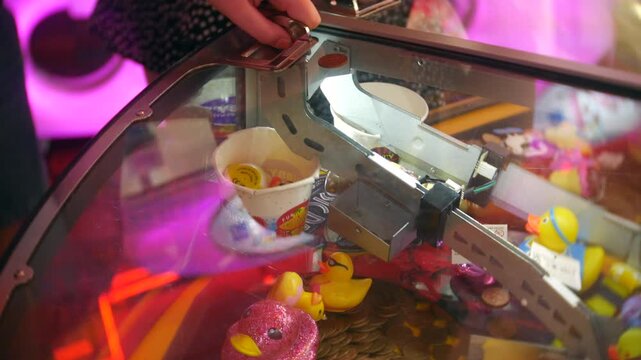 Child operating an arcade claw machine grips the metal control while prizes sit beneath the glass. Close-up view shows hands, bright pink lighting, and colorful toy capsules