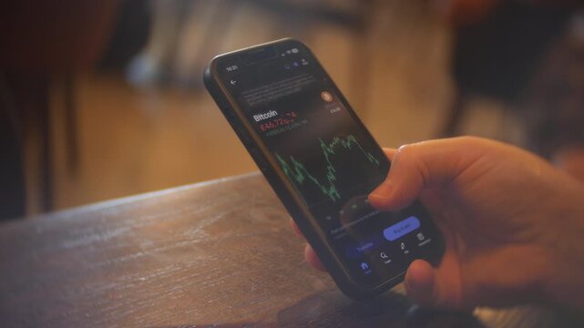 Smartphone displays a Bitcoin price chart as a person holds the device over a wooden table. Close-up view shows cryptocurrency trading data, market graph, and warm indoor light