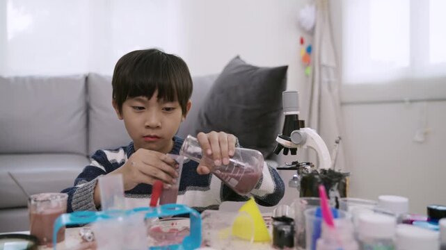 Happy adorable Asian boy with microscope sitting learning science and conducting chemistry experiments by mixing various colored liquids with test tubes and beakers, enjoying in nursery classroom lab.